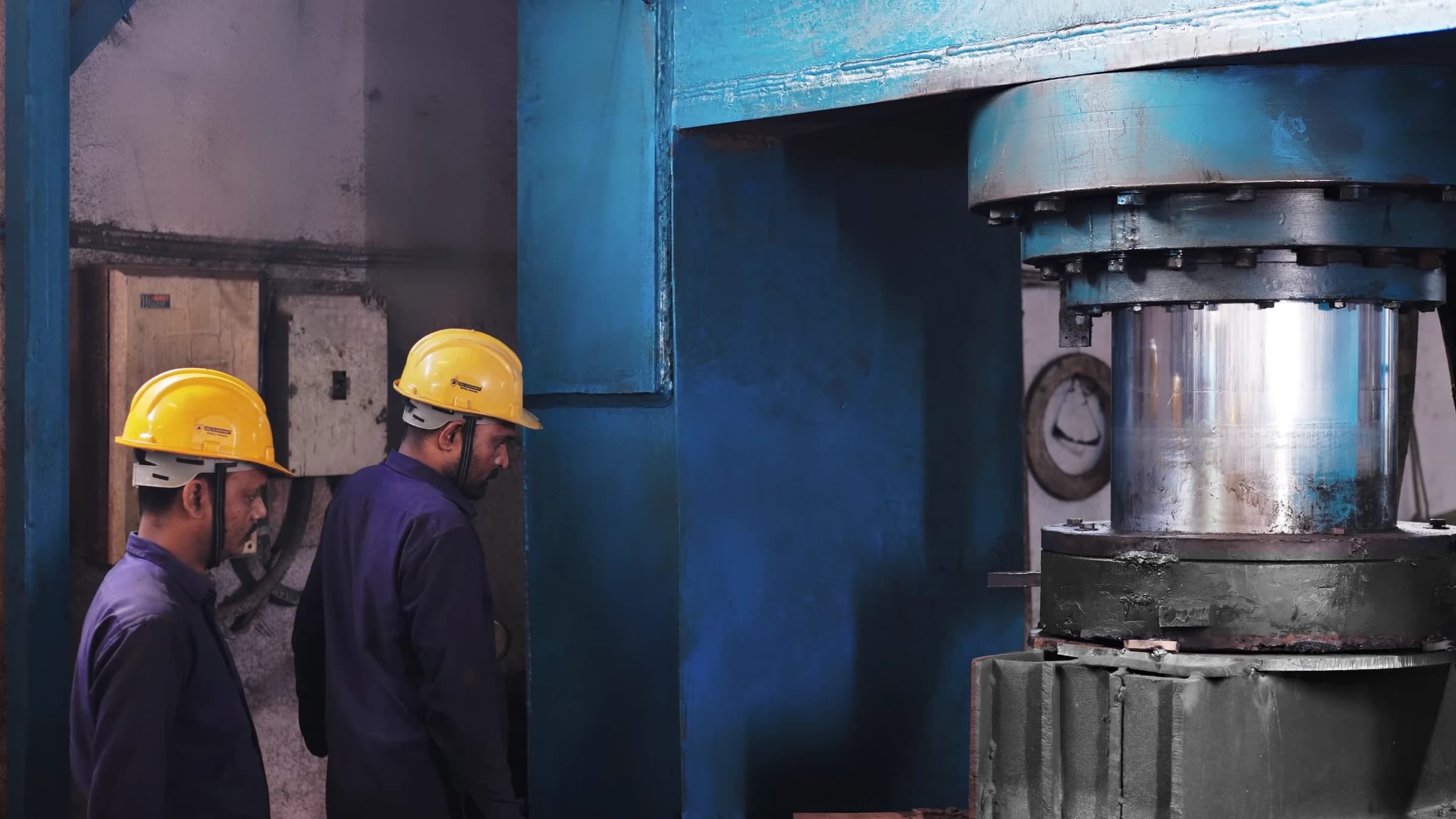 Two operators in safety helmets beside a hydraulic press and its polished ram